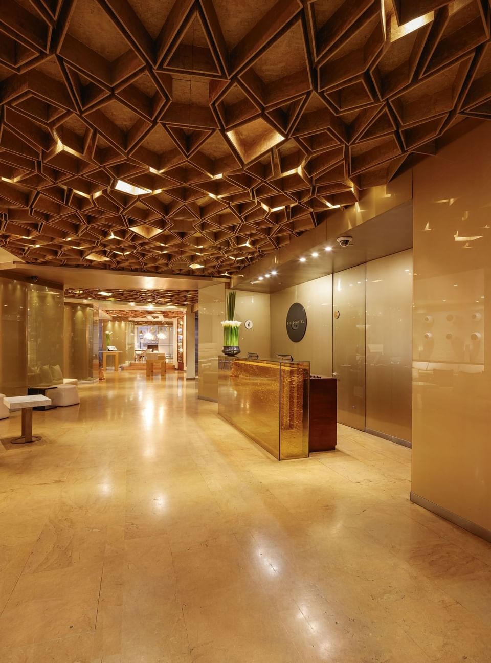 Vestíbulo de hotel con techo geométrico, mostrador de recepción dorado y paredes de vidrio.